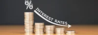 Stacks of coins with a downward arrow labelled interest rates, representing fixed deposit rates, savings returns, and interest-rate movement.