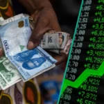 Hands holding Nigerian naira notes beside a stock-market board with a rising green arrow, representing Nigeria’s equity market rally and NGX investor momentum.