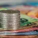 Close-up image of stacked coins on South African rand banknotes with a South African flag icon, representing South African government bonds, rand-denominated fixed income, falling yields and investor focus on bond-market pricing.