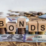 Wooden blocks spelling “Bonds” placed on stacks of coins and U.S. dollar notes, representing Treasury securities, fixed-income investing, bond yields and government-debt markets.