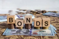Wooden blocks spelling “Bonds” placed on stacks of coins and U.S. dollar notes, representing Treasury securities, fixed-income investing, bond yields and government-debt markets.