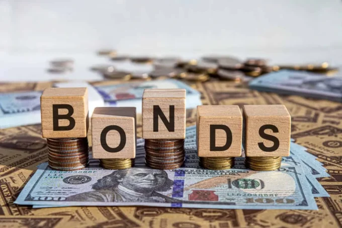 Wooden blocks spelling “Bonds” placed on stacks of coins and U.S. dollar notes, representing Treasury securities, fixed-income investing, bond yields and government-debt markets.