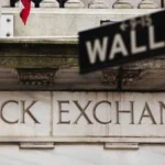 Article image showing the New York Stock Exchange building with a Wall Street sign in the foreground. The image represents U.S. equity markets, Wall Street trading sentiment and investor focus on the S&P 500 near record levels.