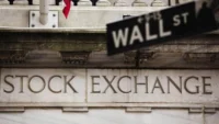 Article image showing the New York Stock Exchange building with a Wall Street sign in the foreground. The image represents U.S. equity markets, Wall Street trading sentiment and investor focus on the S&P 500 near record levels.